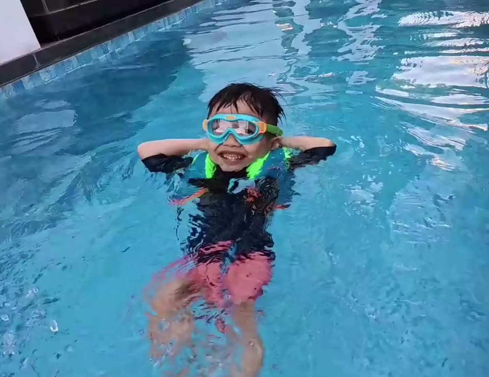 A young child in goggles and a float vest lies back in the water with hands behind their head, grinning.